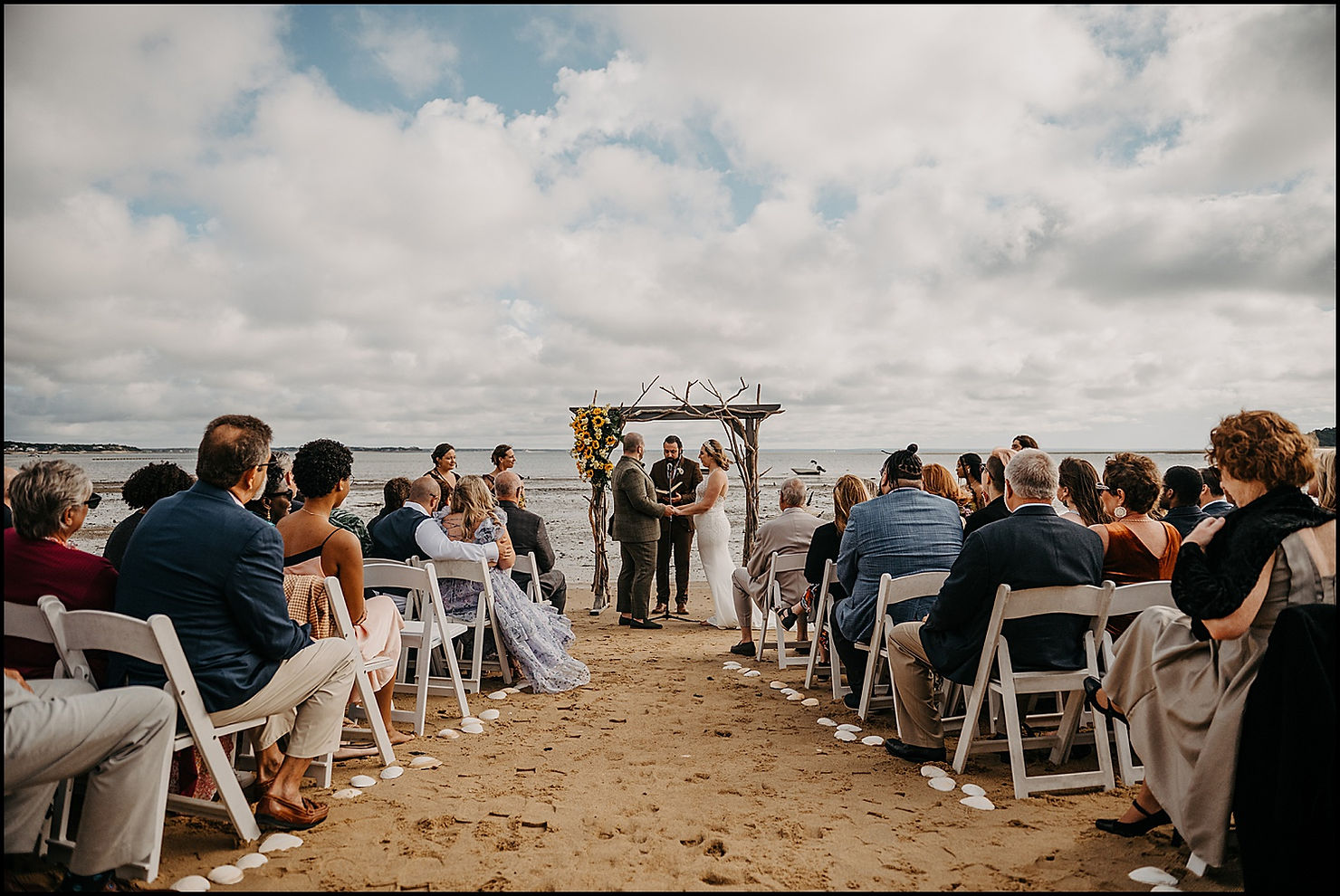 A bride and groom stand beside a driftwood arch for a wedding ceremony on the beach at the Chequessett Club.