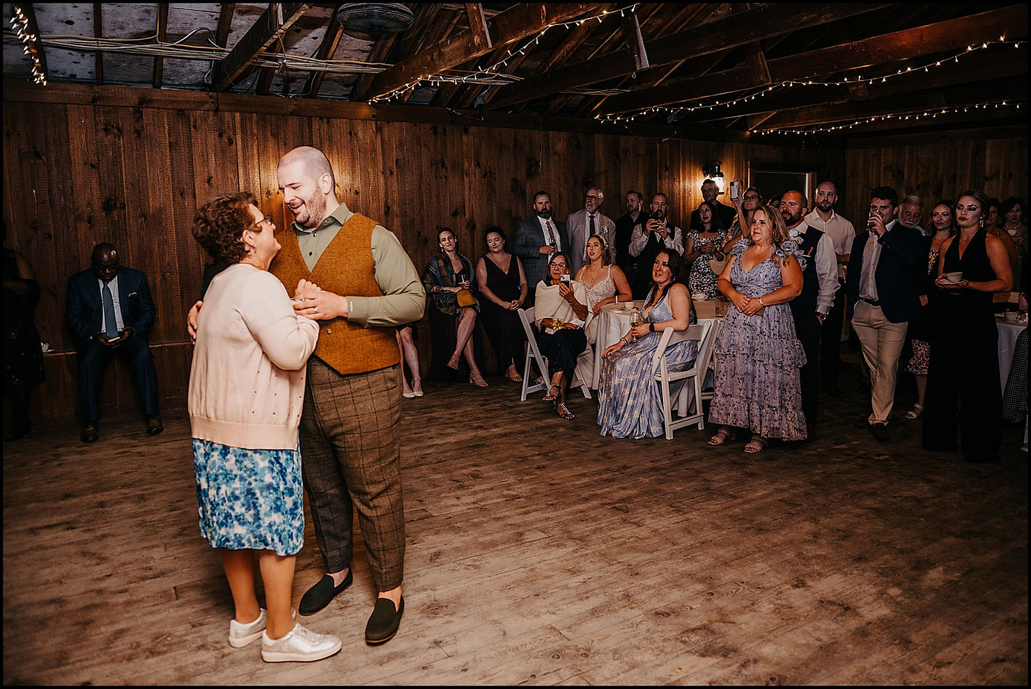 A groom dances with his mother while wedding guests watch from reception tables.