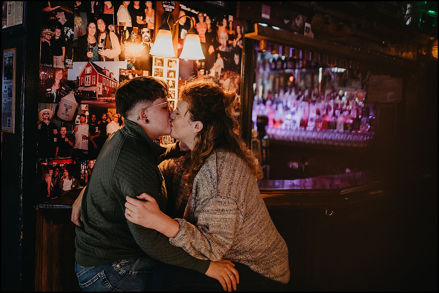 An engaged couple sits in a Buffalo bar and kisses under a sconce.
