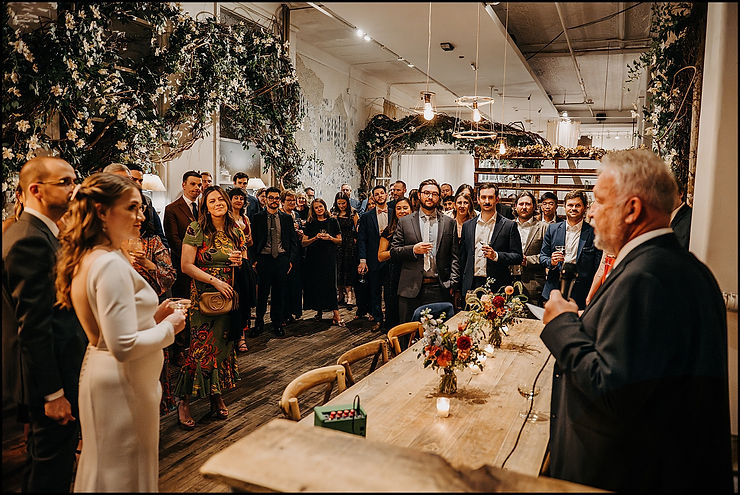 Guests gather around a dining room to listen to a speech at a New York City wedding venue.