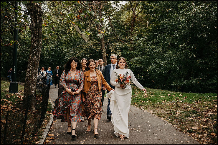 A bride's sister carries her train as they walk down a path towards a wedding in Central Park.